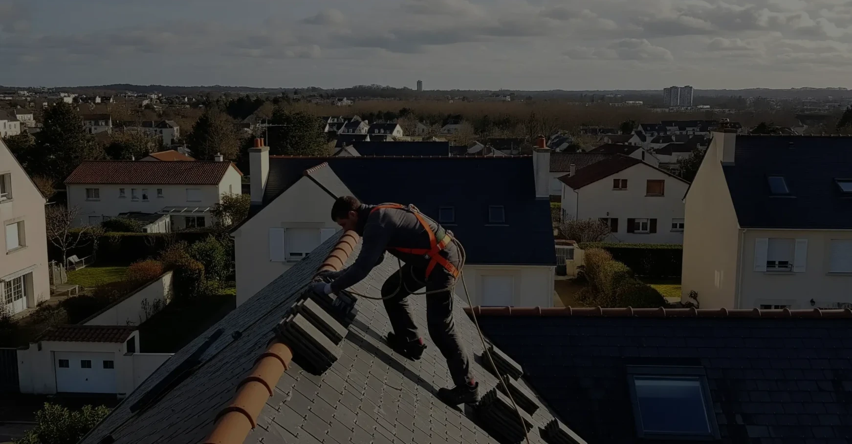 Un couvreur en tenue de travail répare un toit en ardoise sur une maison de la périphérie de Nantes, avec une vue sur la ville en arrière-plan.